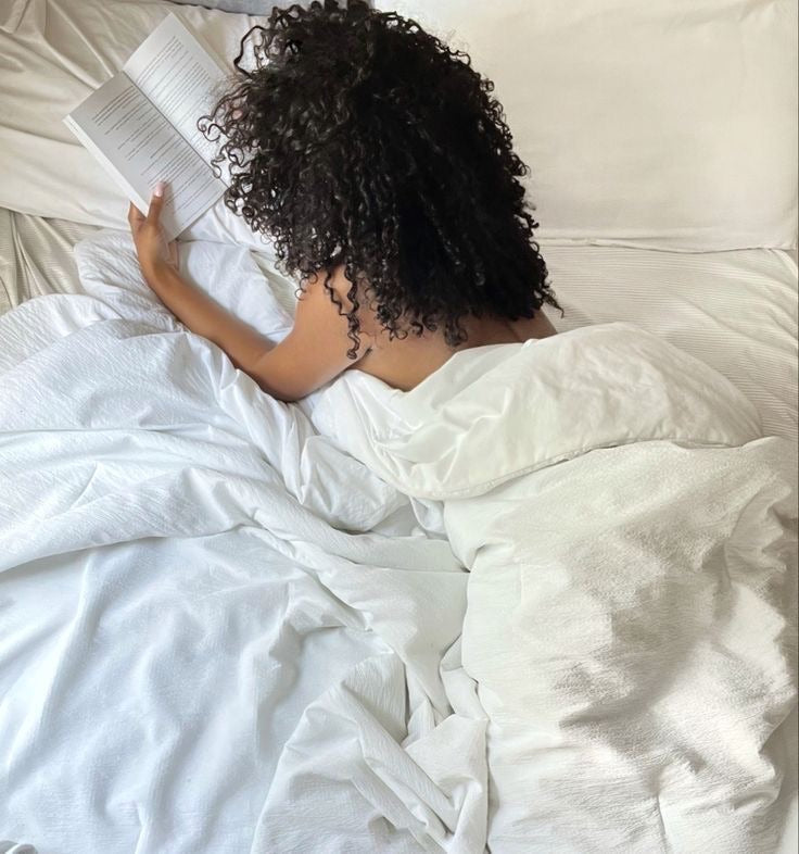 Curly hair girl reading book in bed with white sheets