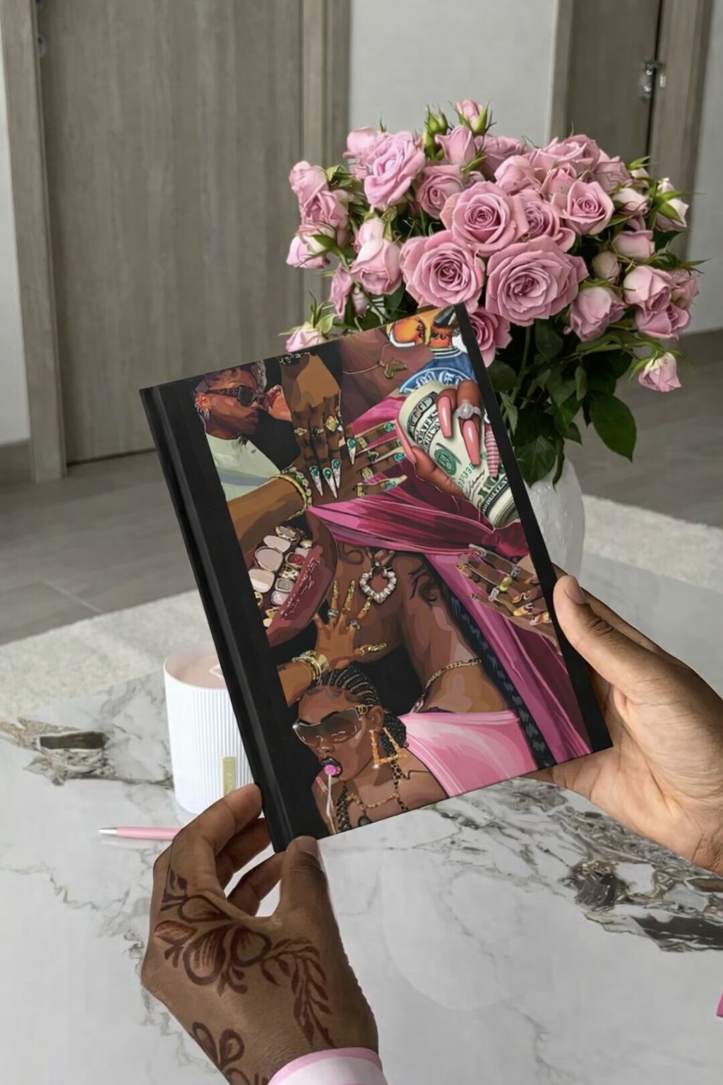Person holding a book with a colorful cover, next to a bouquet of pink roses on a marble surface. Black girl notebook